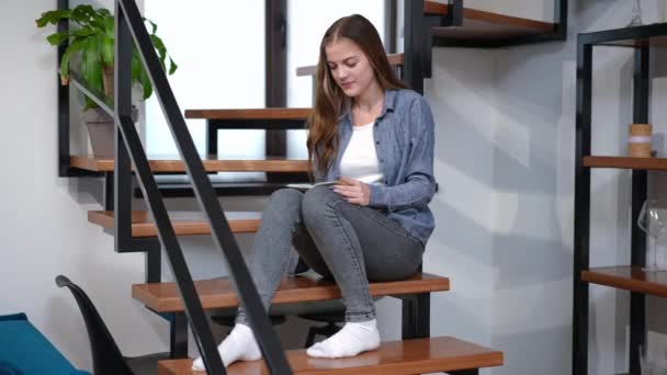 Amplio retrato de una adolescente feliz leyendo un libro de abrazos mirando hacia otro lado soñando. Caucásico bastante adolescente disfrutando de hobby en casa en el interior en fin de semana sentado en escaleras sonriendo. — Vídeos de Stock