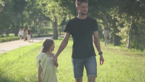 Young Caucasian father and little daughter walking in sunshine in spring summer park holding hands. Portrait of happy man and girl strolling in sunbeam outdoors enjoying family weekend. — Stock Video