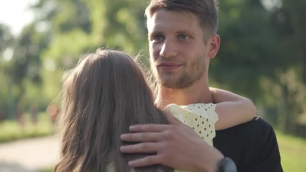 Portret młodego mężczyzny przytulającego dziewczynkę, mówiącego w wiosennym letnim parku w słońcu. Szczęśliwy zrelaksowany ojciec rasy kaukaskiej odpoczywający z córką w weekend na świeżym powietrzu. Wypoczynek i rodzina. — Wideo stockowe