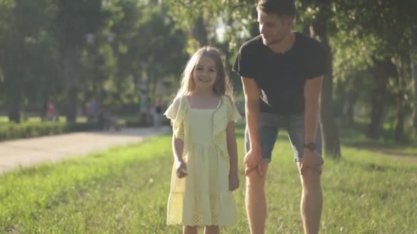 Dolly shot of cheerful cute girl with father in sunny park running looking at camera leaving. Positive Caucasian daughter having fun outdoors in sunshine. Happiness and childhood concept. — Stock Video