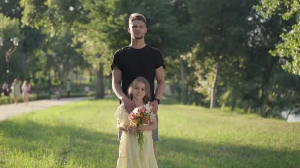 Ευρεία πυροβόλησε μπροστά άποψη αυτοπεποίθηση νεαρός άνδρας και χαριτωμένο γοητευτικό κορίτσι με μπουκέτο λουλούδια κοιτάζοντας κάμερα στέκεται στο ηλιόλουστο πάρκο. Πορτρέτο του ευτυχισμένου Καυκάσου πατέρα ποζάρουν με την κόρη. — Αρχείο Βίντεο