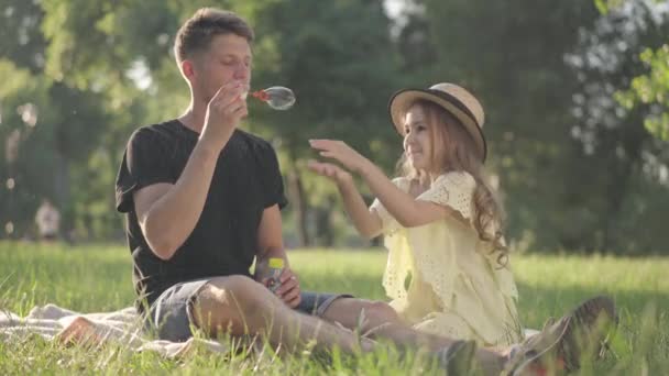 Радостная кавказская дочь ловит мыльные пузыри, когда отец дует мылом, сидя на зеленом весеннем лугу в солнечном парке. Веселый молодой человек и маленькая девочка веселятся, наслаждаясь отдыхом. — стоковое видео