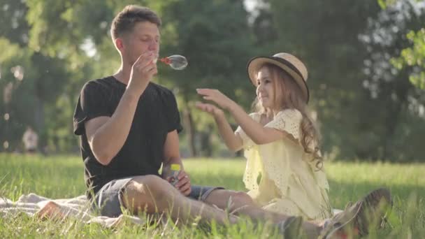 Alegre hija caucásica la captura de burbujas de jabón como padre soplando jabón sud sentado en verde primavera verano prado en el soleado parque. Joven alegre y niña divirtiéndose disfrutando del ocio. — Vídeos de Stock