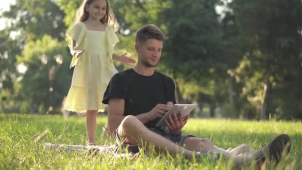 Szeroki strzał zrelaksowany człowiek surfowanie po Internecie na tablecie jako słodka dziewczynka pokrywa oczy rękami. figlarny kaukaski córka odpoczynek z młody przystojny ojciec w słoneczny wiosenny letni park. — Wideo stockowe