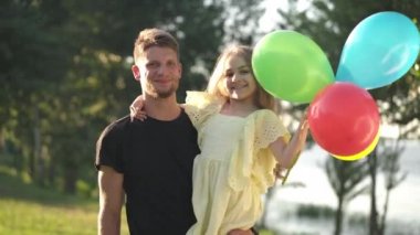 Çok renkli balonlarla kızını kucaklayan ve gülümseyen yakışıklı genç bir baba. Güneşli parkta güneşli bir kızla tatilin tadını çıkaran pozitif beyaz adamın portresi..