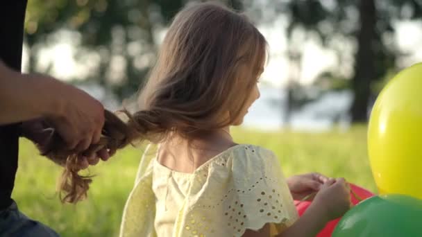 Happy brunette girl playing with balloons as male hands braiding long hair in sunshine. Unrecognizable Caucasian father taking care of little daughter resting in summer spring park outdoors. — Stock Video