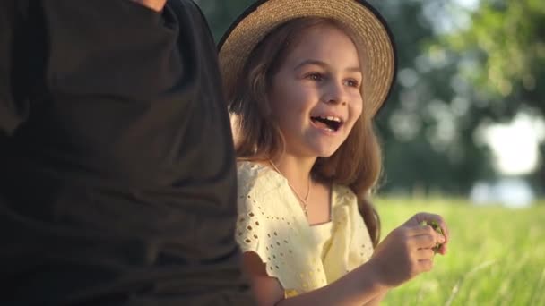 かわいいカリオスの女の子は、認識できない男と緑の春の夏の牧草地の屋外に座って話して笑顔。正白人の娘の肖像とともに父で公園チャット. — ストック動画