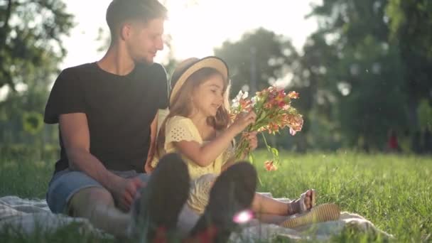 Счастливая расслабленная девушка разговаривает сидя с молодым человеком в сарафане, наслаждаясь семейным уикендом. Портрет очаровательной кавказской дочери с букетом цветов, отдыхающей с отцом на женский день в весеннем парке. — стоковое видео