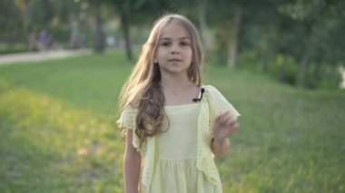 Yaka mikrofonlu sarı elbiseli, kendinden emin beyaz bir kız dışarıda parkta kameraya bakarak konuşuyor. Küçük sevimli blog yazarının portresi podcast 'i çekiyor yaz günü açık havada