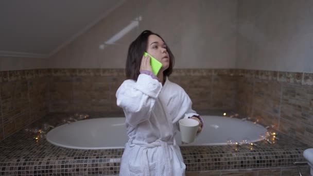 一个自信的小女孩站在家里的浴室里 一边打电话一边喝咖啡 早上穿着白色浴衣的白人小孩的画像 现场摄像机 — 图库视频影像