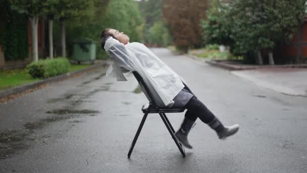 雨のコートを着たリラックスした小さな人と 郊外で曇りの雨の日を楽しむ笑顔の椅子に座っているゴム製のブーツ サイドビュー自信に満ちた白人の小さな女性の広いショット屋外で道に休んで — ストック動画