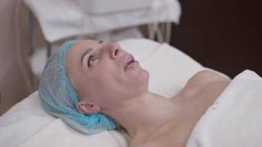 Kendine güvenen, duş şapkalı, spa salonunda uzanmış yavaş çekimde gülümseyen beyaz bir kadın. İçerideki güzellik merkezinde rahatlamanın keyfini çıkaran pozitif, rahat bir kadının portresi.