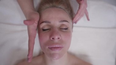 Kafası rahat, beyaz bir kadının kadın elleriyle ağır çekimde yüz masajı yaparken gözlerini kapatması. Çekici bir müşterinin yakın plan portresi güzellik merkezinde masaj yapmaktan zevk alıyor.