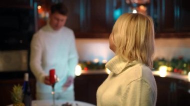 Charming plus-size adult woman looking back at blurred man setting table with romantic dinner turning looking at camera touching chest smiling. Portrait of happy loving wife with husband at home