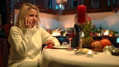 Medium shot portrait of anxious woman sitting at table with romantic dinner talking on phone. Sad Caucasian plus-size girlfriend waiting for partner in kitchen at home. Dating and sadness