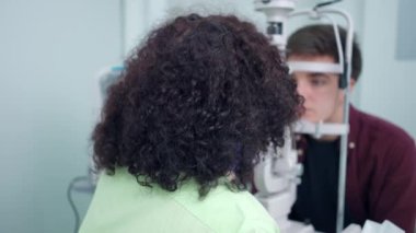 Retina kamerasındaki göz bebeğini kontrol eden profesyonel doktor gülümseyen kameraya bakıyor. Bulanık beyaz adam ekipmanların arkasında oturuyor. Oftalmoloji kavramı