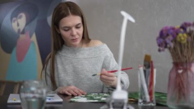 Portrait of charming attractive Caucasian woman painting sitting at table looking out the window smiling. Front view of confident talented beautiful painter enjoying hobby indoors at home