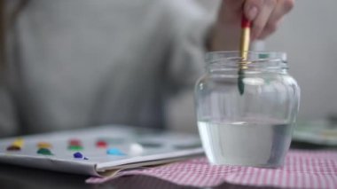 Yakın plan çamaşır fırçası ve şeffaf kavanozda suyun içinde yeşil boya. Tanımlanamayan genç beyaz kadın, kapalı alanda renk ve renk karışımı şeyler çiziyor. Hobi ve ilham perisi kavramı