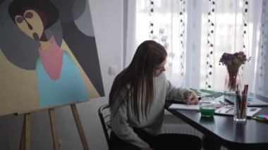 Concentrated young woman stretching hand looking at pencil drawing sketch on paper. Side view portrait of focused talented Caucasian painter creating idea indoors at home. Hobby and talent