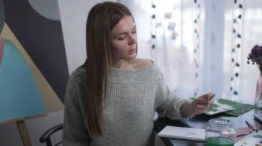 Bir gözünü kapatan kalemle elini uzatıp kameraya gülümseyen neşeli yaratıcı kadın. Kapalı mekanda poz veren neşeli beyaz ressamın portresi. Mutluluk ve hobi