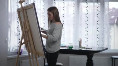 Portrait of talented woman making strokes on easel canvas painting picture indoors. Concentrated attractive Caucasian painter enjoying hobby creating image with acrylic colors. Joy and art