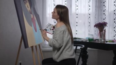 Side view gorgeous female painter making strokes with acrylic paint brush sitting at picture thinking. Talented inspired confident Caucasian woman enjoying hobby indoors. Creativity and fine art