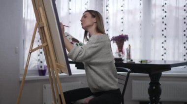 Nilüfer çiçekli kadın sandalyede poz veriyor fırçayla akrilik boya alıyor tuvale dokunuyor. İçeride sanat çalışması yapan yetenekli beyaz bir sanatçıdan esinlendim. İlham perisi ve güven kavramı