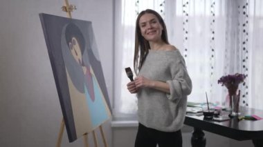 Fotoğraf makinesine gülümseyen genç bir kadının portresi fırça darbeleriyle soyut resim çiziyor. Mutlu yetenekli Kafkasyalı ressam başyapıtla içeride poz veriyor.
