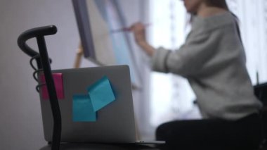Close-up laptop on chair indoors with blurred unrecognizable young painter drawing at background. Slim Caucasian woman sitting at easel with canvas creating picture. Inspiration and muse concept