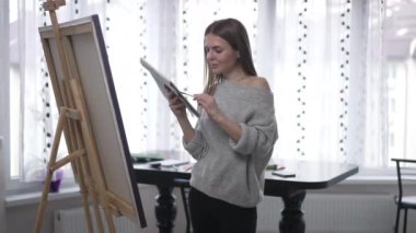 Kendinden emin kadın resim paletiyle poz veriyor. Tabloda, tuvalin içinde duruyor. Kameranın kamerasına gülümseyen beyaz güzel ressama odaklan. Özgüven ve sanat