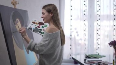 Talented cheerful young woman painting on canvas waving looking away smiling inviting guest. Side view portrait of happy confident Caucasian artist standing at easel creating picture gesturing