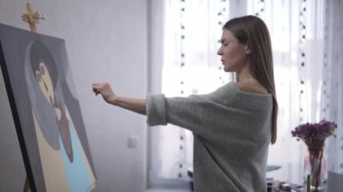 Focused confident woman painting picture stretching hand looking at brush standing at easel. Side view Caucasian concentrated talented painter creating artwork indoors