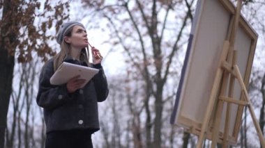 Düşünceli, yaratıcı, güzel bir kadın. Sonbahar bulutlu parkında duruyor. Resim yapmak için uzaklaşıyor. Özümsenmiş beyaz ressam portresi açık havada şaheser yaratıyor.