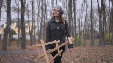 Gülümseyen yetenekli genç bir kadının açık havada yürüdüğünü gösteren bir fotoğraf. Sonbahar parkında resim malzemeleriyle gezip bulutlu havanın tadını çıkaran kendine güvenen Kafkasyalı srtist mutlu.