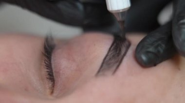 Güzellik uzmanının eliyle yakın plan kadın gözü deriyi pigmentle dolduruyor. Tanımlanamayan profesyonel dövme ustası yüz derisine mikroplu kalem enjekte ediyor.