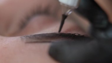 Yakın plan, kaş derisinde mikropatenli kalemle kalıcı mürekkep uygulanıyor. Tanımlanamayan profesyonel güzellik uzmanı. Eldivenlerinde beyaz kadın dövmesi var. İğne ve pigmentle şekil veriyor.