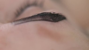 Rack focus along female tattooed eyebrow with hands in gloves appearing with microblading pen. Unrecognizable tattoo master tattooing Caucasian young woman in beauty salon in slow motion