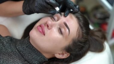 Yüksek açılı beyaz kadın portresi. Kalıcı makyaj yapıyor. Tanınmayan bir güzellik uzmanının kaşına mikropatenli kalem dövmesi yaptırıyor. Güzellik salonunda güzel, ince bir müşteri.