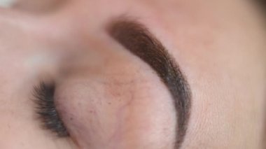 Close-up eye of young Caucasian woman with camera moving to hands in gloves tattooing eyebrow with microblading pen. Unrecognizable expert beautician injecting pigment in facial skin with apparatus