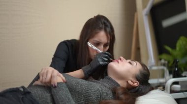 Side view woman lying in beauty salon with blurred concentrated professional beautician contouring upper lip with white in slow motion. Caucasian client and tattoo master doing permanent makeup