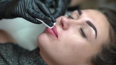 High angle view face of young slim woman in beauty salon with beautician removing blood and red pigment leftovers from lips. Beautiful Caucasian confident client doing permanent lip tattooing