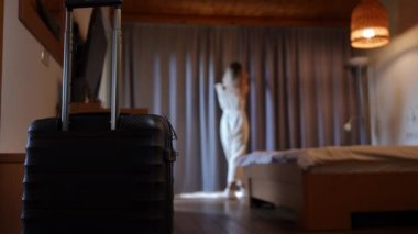 Black suitcase with baggage and blurred Caucasian young woman opening curtains at background in hotel room. Happy tourist with luggage enjoying summer vacations indoors at resort. Slow motion
