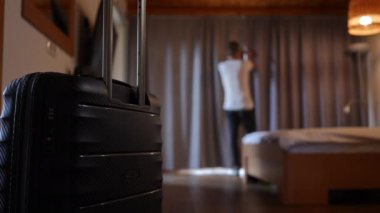 Close-up black travel bag in hotel room with blurred young man opening curtains at background. Confident Caucasian male traveler enjoying summer vacations at cozy resort. Tourism concept
