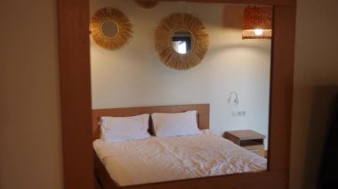 Double bed in hotel room with white clean bedding reflecting in mirror on wall at tourist resort. Comfortable furniture indoors with no people. Tourism and comfort concept
