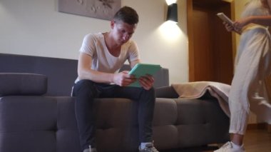 Absorbed young man scrolling social media on tablet as woman entering with book sitting down on couch. Portrait of Caucasian boyfriend messaging online as gilfriend reading on vacations