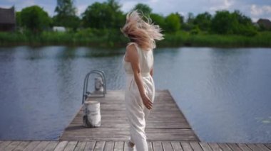Live camera follows joyful smiling young woman running jumping along wooden pier in slow motion posing. Back view tracking shot of cheerful Caucasian beautiful lady enjoying summer leisure at river
