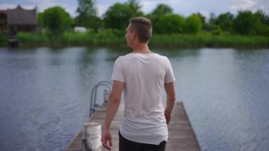 Back view confident young Caucasian man walking in slow motion to river on wooden pier raising arms. Live camera follows fit guy enjoying tranquility outdoors with beautiful landscape at background
