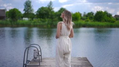 Back view happy confident young woman admiring beauty of nature standing on wooden pier at blue river. Relaxed carefree Caucasian lady smiling looking away in slow motion. Life balance concept