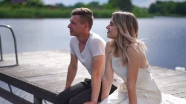 Young romantic Caucasian couple sitting on wooden pier talking dating on sunny summer day outdoors. Portrait of happy loving man and woman enjoying leisure at river in slow motion