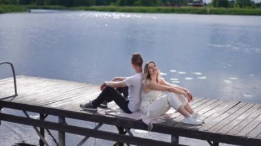 Side view wide shot relaxed Caucasian young couple sitting back to back on wooden pier with river water in sunshine at background. Happy carefree man and woman talking laughing in slow motion